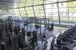 Ein großzügiges, modernes Fitnessstudio mit großen, bodentiefen Fenstern, durch die viel Tageslicht einfällt. Verschiedene Trainingsgeräte wie Laufbänder und Kraftstationen sind ordentlich im Raum verteilt. Einige Personen trainieren—eine Person hebt Gewichte an einem funktionellen Trainingsgerüst, während andere Cardiogeräte nutzen. Die Umgebung wirkt sauber, hell und gut organisiert.