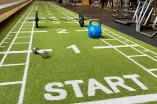 In einem modernen Fitnessbereich liegt eine grüne Sprintbahn mit weißen Markierungen und dem Wort „START“ am Anfang. Entlang der Bahn sind verschiedene Trainingsgeräte wie eine Hantel, eine Kettlebell und eine Langhantel verteilt. Im Hintergrund sieht man ein lichtdurchflutetes Studio mit offenen Trainingsbereichen und motivierenden Schriftzügen wie „THINK.MOVE. LIVE.“ an der Wand. Die Atmosphäre wirkt einladend und aktivitätsfördernd.