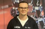 Eine Person steht vor einem Hintergrund mit unscharfen Hanteln und Fitnessgeräten. Die Person trägt eine dunkle Kapuzenjacke mit mehreren Logos und ein Namensschild. Sie hat kurze Haare und eine runde Brille. Die Person lächelt leicht in die Kamera und wirkt in einer professionellen, sportbezogenen Umgebung.