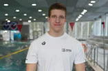 Eine Person steht in einer Schwimmhalle vor einem Becken mit Schwimmbahnen. Sie trägt ein weißes Sport-T-Shirt und lächelt leicht in die Kamera. Im Hintergrund sind Wasser, Leinen, Fenster und Hallenbeleuchtung zu sehen.