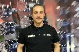 Eine Person steht in einem Fitnessstudio vor einem Hantelregal. Sie trägt ein schwarzes Shirt und eine helle Hose, eine Hand ruht auf der Hüfte. Der Hintergrund ist leicht unscharf, wodurch die Gewichte betont werden und eine räumliche Tiefe entsteht.
