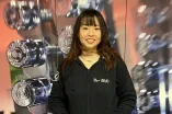 Eine Person steht vor einer Wand mit mehreren glänzenden Hanteln. Sie trägt einen schwarzen Hoodie mit „RUB“-Logo und einen dunklen Rock. Die Hände sind in den Taschen, und sie lächelt in die Kamera. Das Foto wirkt wie in einem Fitnessstudio aufgenommen.
