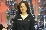 ine Person steht vor einer Wand mit vielen glänzenden Hanteln im Hintergrund. Sie trägt einen schwarzen Hoodie mit Fitnessstudio-Logo und lächelt leicht in die Kamera. Die Hände sind vor dem Körper gefaltet, und das Bild wirkt wie in einem Fitnessstudio oder Trainingsbereich aufgenommen.