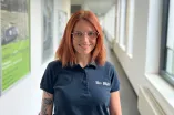 Eine Person mit schulterlangen roten Haaren und Brille steht lächelnd in einem hellen Flur. Die Person trägt ein dunkles Poloshirt mit RUB-Logo und hat sichtbare Tattoos am linken Arm. Im Hintergrund befinden sich Fenster und gerahmte Poster an der Wand.
