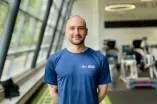 Eine Person mit Kurzhaarschnitt steht lächelnd in einem modernen Fitnessstudio. Sie trägt ein dunkelblaues T-Shirt mit dem RUB-Logo auf der Brust. Im Hintergrund sind Trainingsgeräte und große Fenster zu sehen.