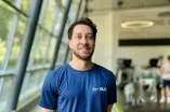 Eine Person mit kurzem dunklen Haar steht lächelnd in einem modernen Fitnessstudio. Sie trägt ein dunkelblaues T-Shirt mit dem RUB-Logo auf der Brust. Im Hintergrund sind Trainingsgeräte und große Fenster zu sehen.