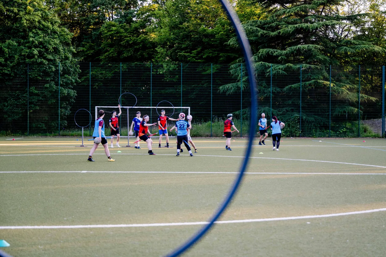 The image shows a group of people on a sports field playing quadball. In the background, there are three goal frames with rings at different heights. A large blue ring is partially visible in the foreground. The area is surrounded by trees.