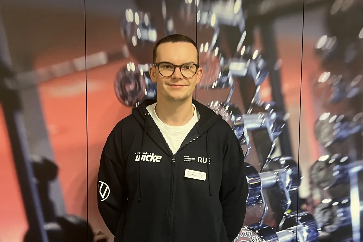 Eine Person steht vor einem Hintergrund mit unscharfen Hanteln und Fitnessgeräten. Die Person trägt eine dunkle Kapuzenjacke mit mehreren Logos und ein Namensschild. Sie hat kurze Haare und eine runde Brille. Die Person lächelt leicht in die Kamera und wirkt in einer professionellen, sportbezogenen Umgebung.