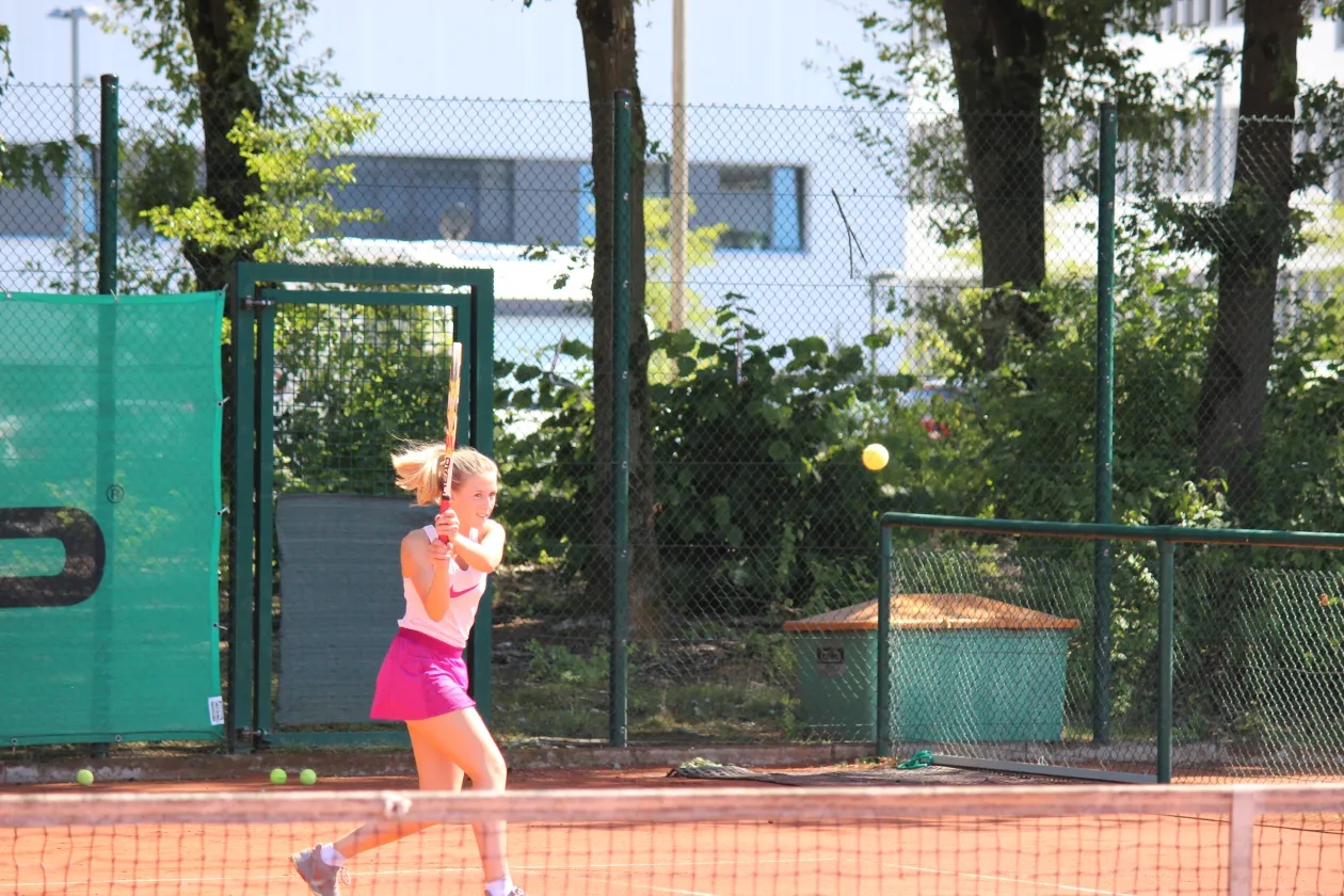 A woman is playing tennis on an outdoor court. She is holding her racket up and has the ball in front of her. She takes a step toward the ball.