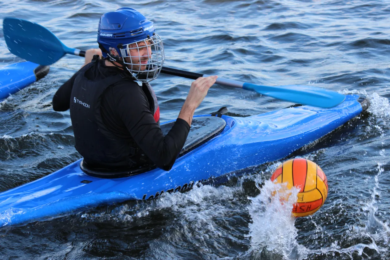 Ein Mann sitzt in einem blauen Kajak auf dem Wasser, trägt einen Helm mit Gesichtsgitter und hält ein Paddel in der Hand. Vor ihm spritzt ein gelb-roter Ball aus dem Wasser, was auf ein sportliches Spiel hindeutet.