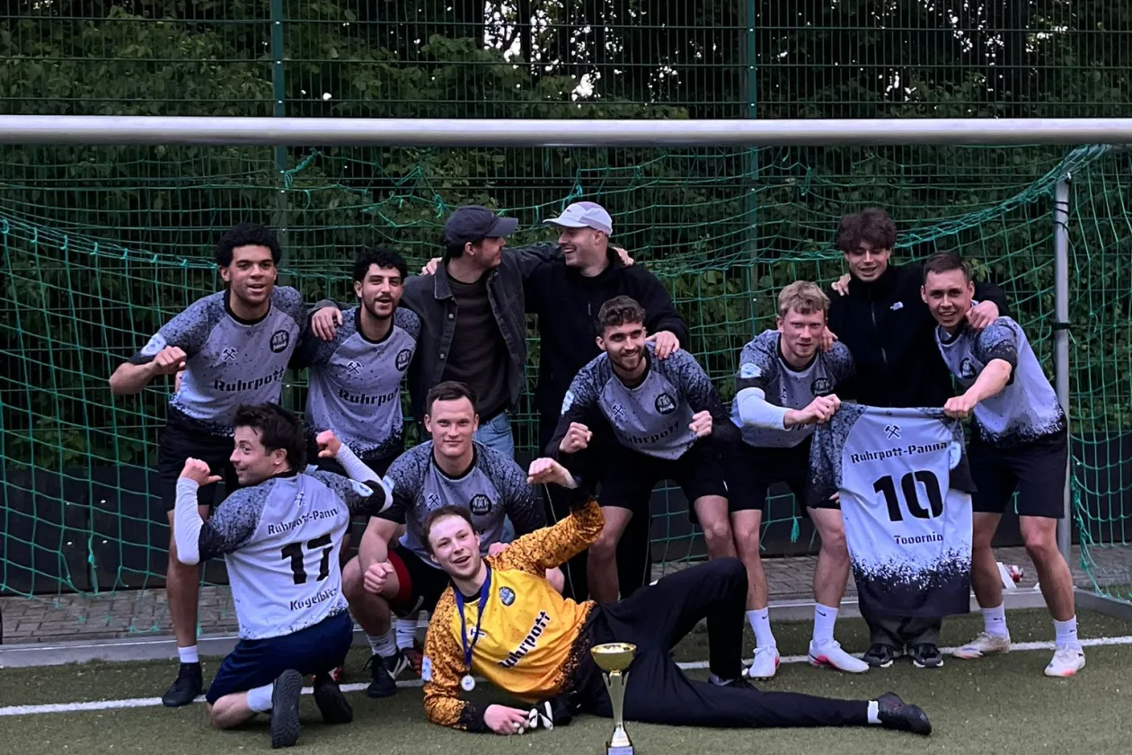 The Ruhrpott-PANNA team is celebrating wildly on the artificial turf field at Bochum University Sports after their victory in the final. Holding aloft the trophy, medals, and a jersey, the team proudly poses after a perfect season and winning the championship.