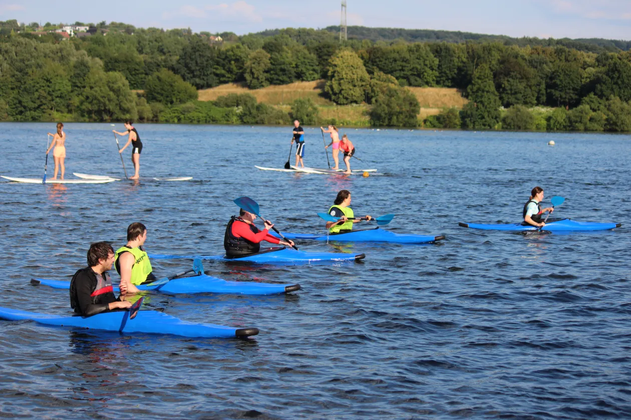 Es paddeln fünf Personen auf dem See in einem Kanu und im Hintergrund stehen weitere 5 Personen auf einem Stand Up Paddle Brett.