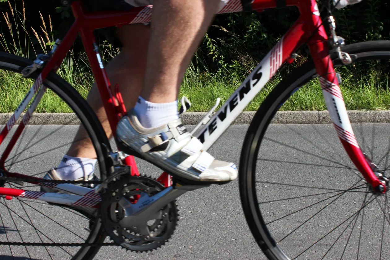 Ein Radfahrer fährt auf einem roten Rennrad auf einer asphaltierten Straße. Sichtbar sind die Beine, die Fahrradschuhe sowie Teile des Fahrrads; der Hintergrund zeigt einen begrünten Straßenrand.