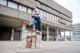 Die Spitzensportlerin Felicitas Merker springt über einen großen Stapel aus Büchern, welche draußen vor einem Unigebäude aufgebaut wurden.