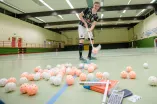 Der Floorballspieler Jan Niklar Buckermann steht in einer Sporthalle und schlägt einen Ball mit seinem Floorballschläger. Vor ihm liegt ein STapel aus Bällen, Büchern und einem Taschenrechner.