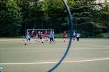 The image shows a group of people on a sports field playing quadball. In the background, there are three goal frames with rings at different heights. A large blue ring is partially visible in the foreground. The area is surrounded by trees.