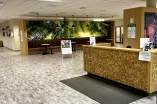 The image shows the reception area of a fitness or wellness center called "UNIfit," with the slogan “get in motion.” The reception desk has a stylish wooden design made of square blocks, and a small mat lies in front of it. Behind the desk are signs and information sheets. To the left, there is a cozy seating area with red stools, small round tables, and a long cushioned bench, all placed against a large forest-themed wall mural that brings a natural and calming atmosphere. Pendant lights hang above the sea