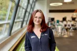 Eine Person mit schulterlangem rötlichem Haar steht in einem Fitnessstudio auf einem grünen Bodenstreifen mit weißen Markierungen. Die Person trägt eine dunkle Kapuzenjacke und graue Sportleggings, hat die Hände in den Jackentaschen und lächelt leicht in die Kamera. Im Hintergrund sind Trainingsgeräte und große Fenster zu sehen, durch die Tageslicht einfällt.