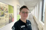A person with short hair and glasses is standing in a bright hallway. The person is wearing a black polo shirt with logos and is smiling at the camera. Windows line the right side of the hallway, and information boards are visible on the wall.