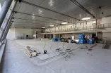 Eine große, leere Innenhalle, die sich offenbar im Umbau befindet. Der Boden ist frei von Wasser, und an mehreren Stellen liegen Baumaterialien wie Holzleisten, Metallstangen und Werkzeuge. Einige Poolleitern sind ausgebaut und liegen auf dem Boden. Im Hintergrund sieht man eine erhöhte Galerie mit Geländer sowie große Fenster, die viel Licht in den Raum lassen. Die Umgebung wirkt unfertig und industriell.