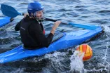 A man is sitting in a blue kayak on the water, wearing a helmet with a face guard and holding a paddle. In front of him, a yellow-and-red ball is splashing out of the water, suggesting a sports game.