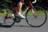 The photo shows a person riding a red racing bike on a paved road. The narrow tires, the hunched riding position, and the curved handlebars are typical features of a racing bike. In the background, there are green spaces and vegetation, suggesting a route through a natural setting.