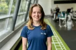 Porträt von Leah Malek in einem hellen, modernen Fitnessstudio. Leah trägt ein blaues Sportshirt mit dem Logo der Ruhr-Universität Bochum und blickt direkt in die Kamera. Im Hintergrund sind Trainingsgeräte und eine große Fensterfront zu sehen.