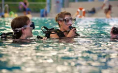 Zwei Personen mit Taucherbrillen und Atemreglern üben in einem Hallenbad. Sie sind teilweise im Wasser und scheinen einer weiteren Person außerhalb des Bildausschnitts zuzuhören oder mit ihr zu sprechen. Der Poolbereich ist hell beleuchtet, und im Hintergrund sind weitere Personen sowie Ausrüstung zu erkennen.