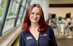 A person with shoulder-length reddish hair is standing in a gym on a green floor track with white markings. The person is wearing a dark hoodie and gray workout leggings, has their hands in the hoodie pockets, and is smiling slightly at the camera. In the background, gym equipment and large windows letting in daylight are visible.