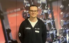 Eine Person steht vor einem Hintergrund mit unscharfen Hanteln und Fitnessgeräten. Die Person trägt eine dunkle Kapuzenjacke mit mehreren Logos und ein Namensschild. Sie hat kurze Haare und eine runde Brille. Die Person lächelt leicht in die Kamera und wirkt in einer professionellen, sportbezogenen Umgebung.