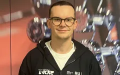 Eine Person steht vor einem Hintergrund mit unscharfen Hanteln und Fitnessgeräten. Die Person trägt eine dunkle Kapuzenjacke mit mehreren Logos und ein Namensschild. Sie hat kurze Haare und eine runde Brille. Die Person lächelt leicht in die Kamera und wirkt in einer professionellen, sportbezogenen Umgebung.