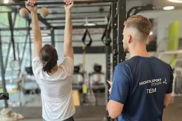 Eine Person hängt sich an eine Klimmzugstange, während eine zweite Person daneben steht und das Training beobachtet oder begleitet. Beide befinden sich in einem modernen Fitnessstudio mit verschiedenen Trainingsgeräten im Hintergrund. Die stehende Person trägt ein dunkelblaues T-Shirt mit der Aufschrift „HOCHSCHULSPORT BOCHUM – TEAM“ auf dem Rücken.
