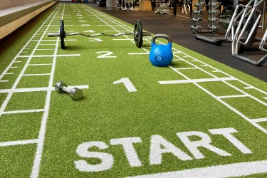 In einem modernen Fitnessbereich liegt eine grüne Sprintbahn mit weißen Markierungen und dem Wort „START“ am Anfang. Entlang der Bahn sind verschiedene Trainingsgeräte wie eine Hantel, eine Kettlebell und eine Langhantel verteilt. Im Hintergrund sieht man ein lichtdurchflutetes Studio mit offenen Trainingsbereichen und motivierenden Schriftzügen wie „THINK.MOVE. LIVE.“ an der Wand. Die Atmosphäre wirkt einladend und aktivitätsfördernd.