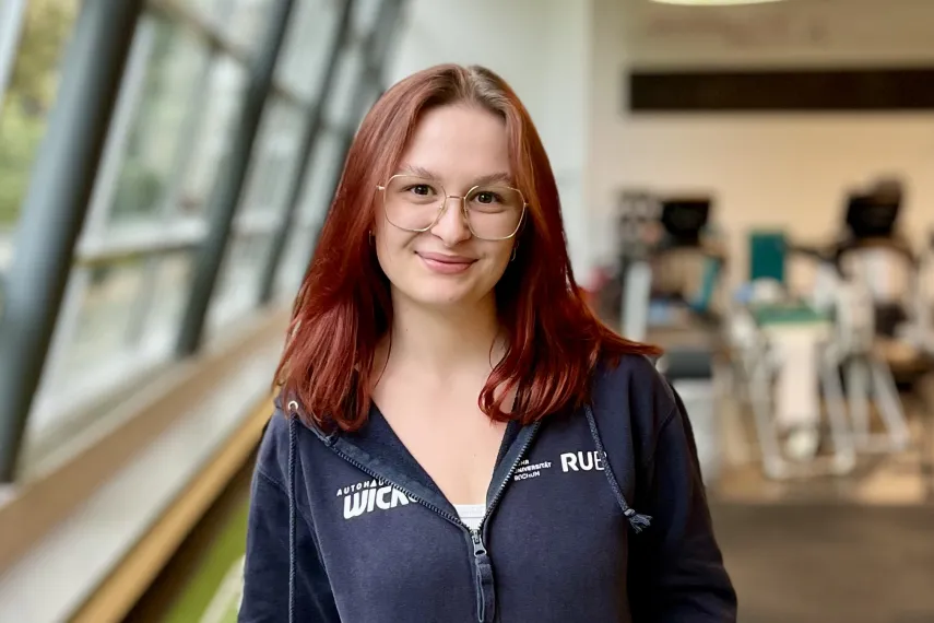 A person with shoulder-length reddish hair is standing in a gym on a green floor track with white markings. The person is wearing a dark hoodie and gray workout leggings, has their hands in the hoodie pockets, and is smiling slightly at the camera. In the background, gym equipment and large windows letting in daylight are visible.