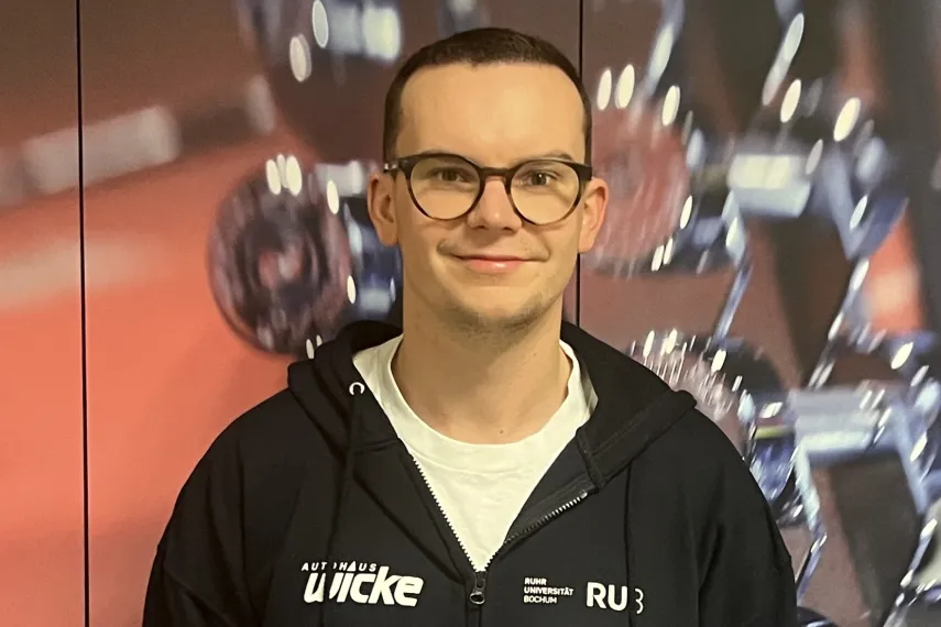 Eine Person steht vor einem Hintergrund mit unscharfen Hanteln und Fitnessgeräten. Die Person trägt eine dunkle Kapuzenjacke mit mehreren Logos und ein Namensschild. Sie hat kurze Haare und eine runde Brille. Die Person lächelt leicht in die Kamera und wirkt in einer professionellen, sportbezogenen Umgebung.