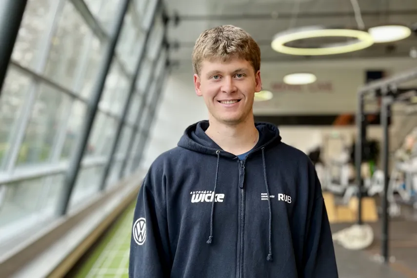 Eine Person steht in einem modernen Fitnessstudio und blickt lächelnd in die Kamera. Sie trägt einen dunklen Kapuzenpullover mit Reißverschluss und eine dunkle Sporthose. Im Hintergrund sind große Fenster, Trainingsgeräte und eine grüne Laufbahn auf dem Boden zu sehen. Die Atmosphäre wirkt hell und sportlich.