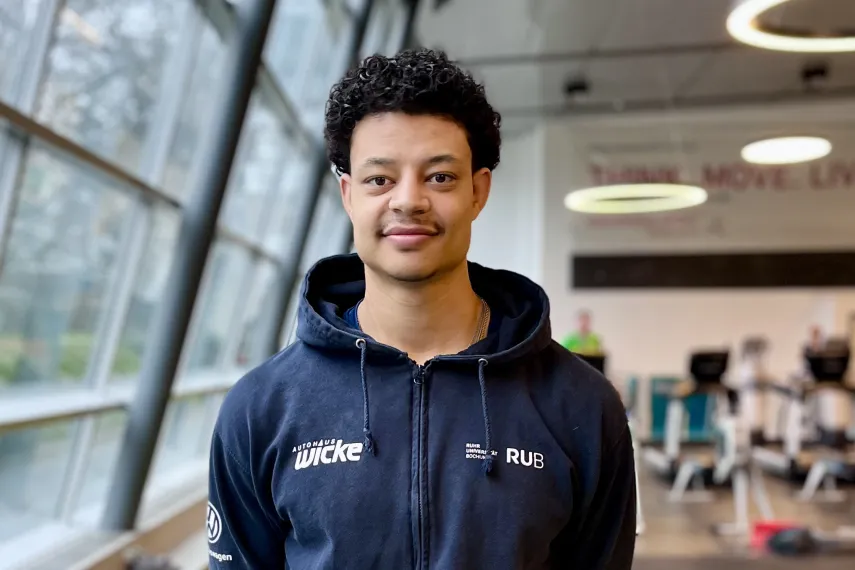 Eine Person mit kurzen, lockigen Haaren steht entspannt in einem modernen Raum. Sie trägt einen dunklen Hoodie und hält die Hände hinter dem Rücken. Durch die großen Fenster fällt Tageslicht ein, im Hintergrund sind Sport- oder Trainingsgeräte zu sehen.