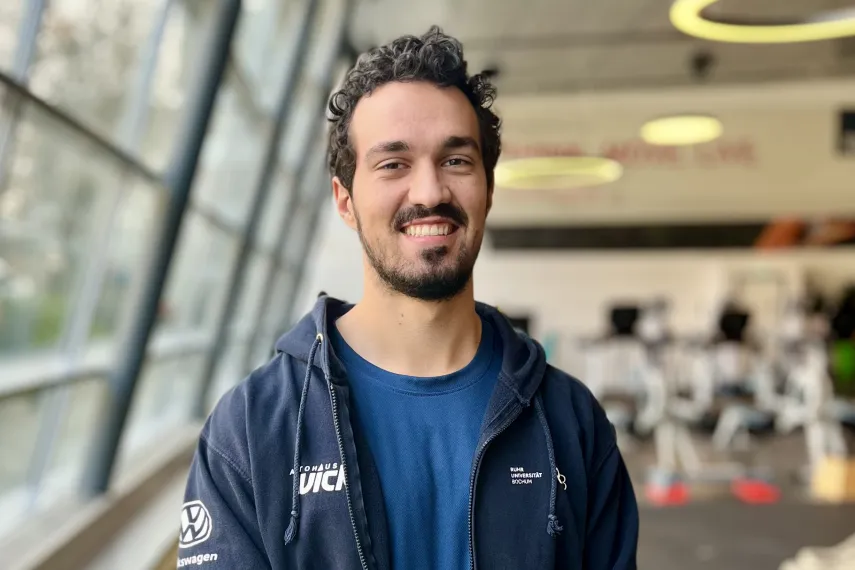 Eine Person mit lockigem Haar steht lächelnd in einem modernen Fitnessstudio. Sie trägt ein dunkles Sweatshirt über einem T-Shirt. Im Hintergrund sind Trainingsgeräte und große Fenster zu sehen.