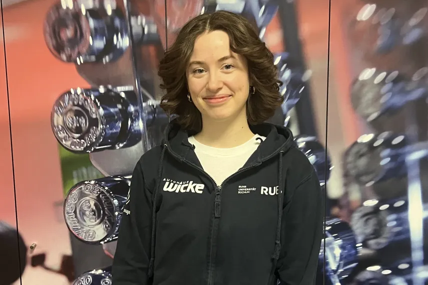 ine Person steht vor einer Wand mit vielen glänzenden Hanteln im Hintergrund. Sie trägt einen schwarzen Hoodie mit Fitnessstudio-Logo und lächelt leicht in die Kamera. Die Hände sind vor dem Körper gefaltet, und das Bild wirkt wie in einem Fitnessstudio oder Trainingsbereich aufgenommen.