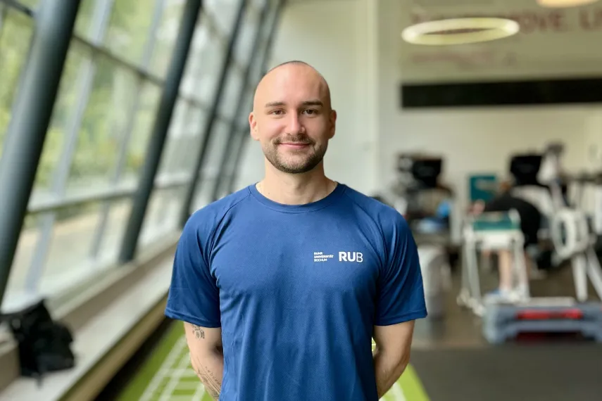 Eine Person mit Kurzhaarschnitt steht lächelnd in einem modernen Fitnessstudio. Sie trägt ein dunkelblaues T-Shirt mit dem RUB-Logo auf der Brust. Im Hintergrund sind Trainingsgeräte und große Fenster zu sehen.