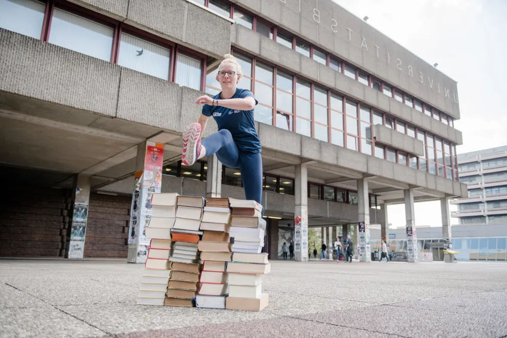 Die Spitzensportlerin Felicitas Merker springt über einen großen Stapel aus Büchern, welche draußen vor einem Unigebäude aufgebaut wurden.