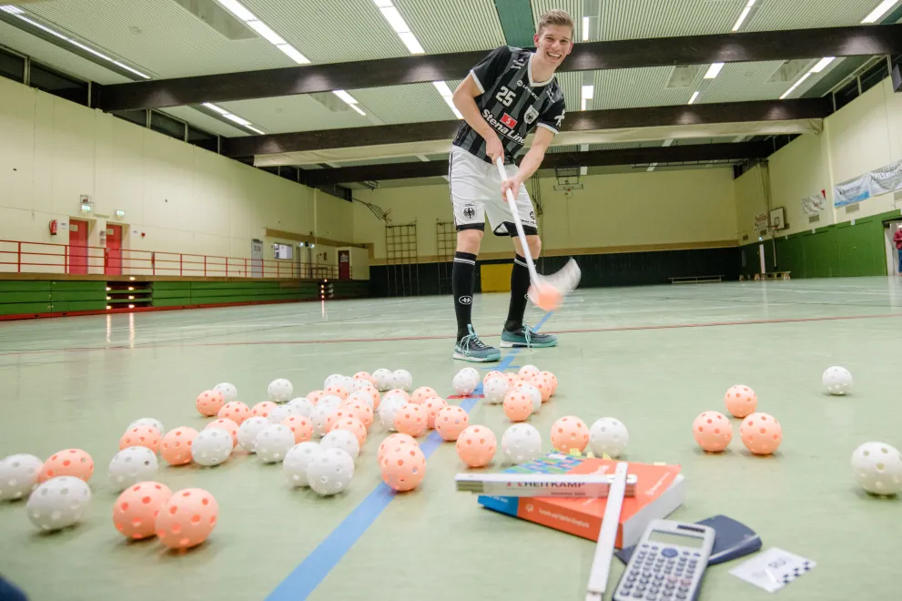 Der Floorballspieler Jan Niklar Buckermann steht in einer Sporthalle und schlägt einen Ball mit seinem Floorballschläger. Vor ihm liegt ein STapel aus Bällen, Büchern und einem Taschenrechner.
