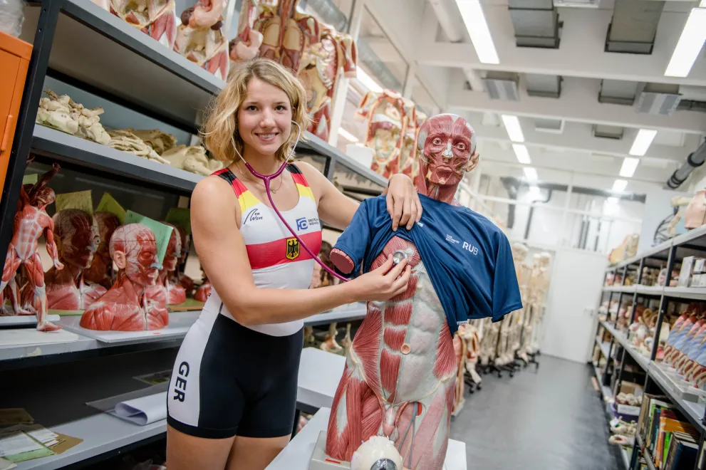 Die Spitzensportlerin Charlotte Reinhardt hört mit einem Sthetoskop eine Puppe ab. Sie trägt dabei einen Rudereinteiler.