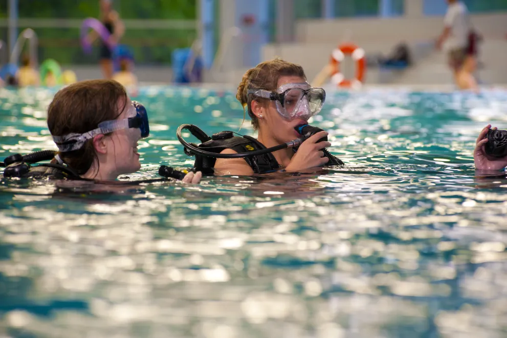 Zwei Personen mit Taucherbrillen und Atemreglern üben in einem Hallenbad. Sie sind teilweise im Wasser und scheinen einer weiteren Person außerhalb des Bildausschnitts zuzuhören oder mit ihr zu sprechen. Der Poolbereich ist hell beleuchtet, und im Hintergrund sind weitere Personen sowie Ausrüstung zu erkennen.