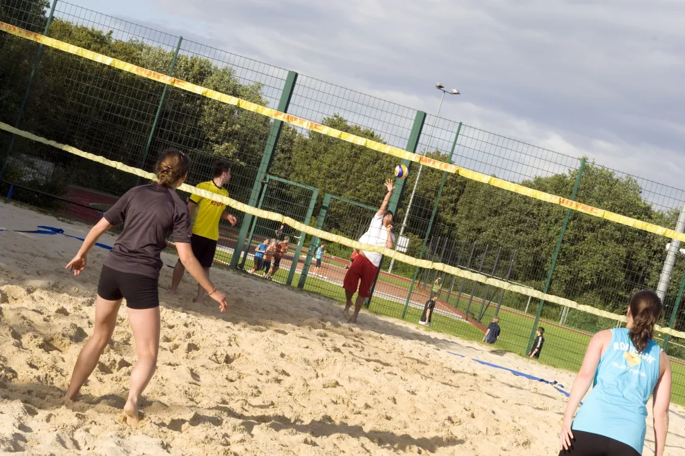 Mehrere Personen spielen Beachvolleyball auf einem Sandfeld. Im Vordergrund sind drei Spielende zu sehen, die aktiv am Spielgeschehen teilnehmen. Der Ball befindet sich in der Luft, während sich die Beteiligten auf beiden Seiten des Netzes positionieren. Im Hintergrund sind grüne Bäume und ein teilweise bewölkter Himmel zu erkennen. Die Szene vermittelt eine sportliche und dynamische Atmosphäre.