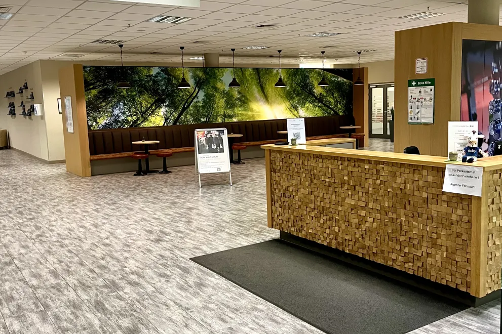 Das Bild zeigt den Empfangsbereich eines Fitness- oder Gesundheitszentrums namens "UNIfit" mit dem Slogan „get in motion“. Der Empfangstresen besteht aus stilvoll angeordneten Holzklötzchen, davor liegt eine kleine Matte. Auf dem Tresen befinden sich Informationsblätter und Aushänge. Links davon ist eine gemütliche Sitzecke mit roten Hockern, kleinen runden Tischen und einer langen gepolsterten Sitzbank, die vor einer großen Wand mit einem beruhigenden Waldmotiv steht. Hängelampen sorgen für angenehmes Lich