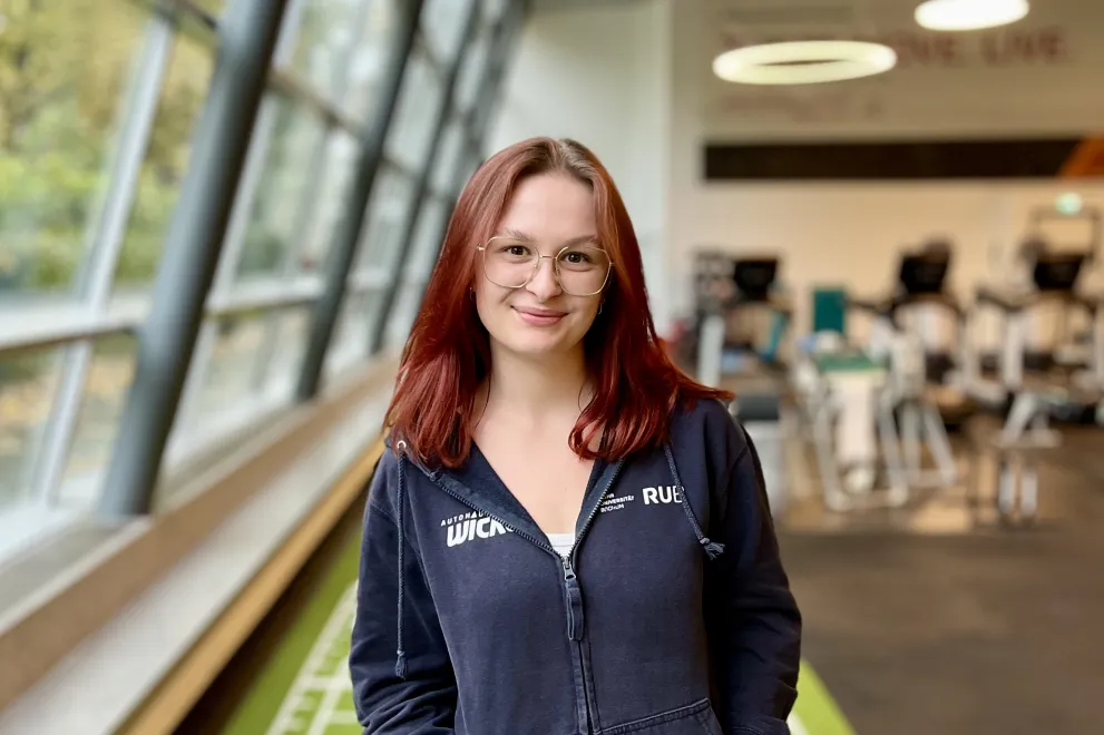 Eine Person mit schulterlangem rötlichem Haar steht in einem Fitnessstudio auf einem grünen Bodenstreifen mit weißen Markierungen. Die Person trägt eine dunkle Kapuzenjacke und graue Sportleggings, hat die Hände in den Jackentaschen und lächelt leicht in die Kamera. Im Hintergrund sind Trainingsgeräte und große Fenster zu sehen, durch die Tageslicht einfällt.