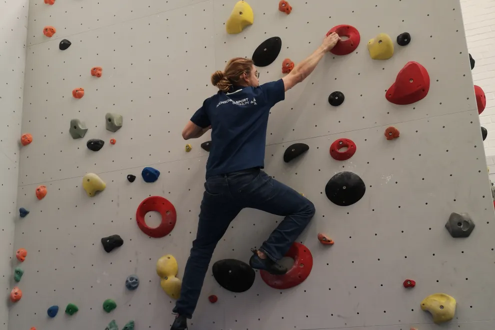 A person wearing a blue T-shirt and jeans is climbing on an indoor bouldering wall. They are reaching upward with one hand toward a red hold while standing on several black and red footholds. The wall is covered with holds of different shapes and colors, and the person appears focused and balanced as they move upward.