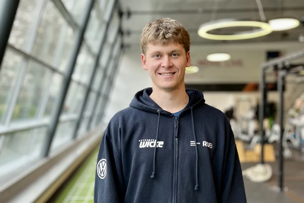 Eine Person steht in einem modernen Fitnessstudio und blickt lächelnd in die Kamera. Sie trägt einen dunklen Kapuzenpullover mit Reißverschluss und eine dunkle Sporthose. Im Hintergrund sind große Fenster, Trainingsgeräte und eine grüne Laufbahn auf dem Boden zu sehen. Die Atmosphäre wirkt hell und sportlich.