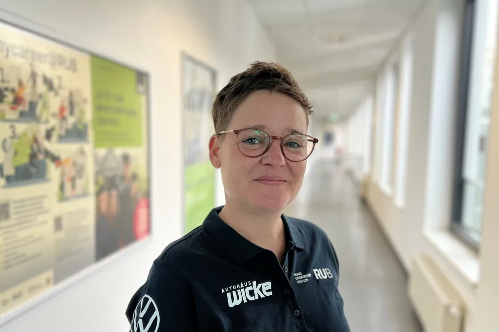 A person with short hair and glasses is standing in a bright hallway. The person is wearing a black polo shirt with logos and is smiling at the camera. Windows line the right side of the hallway, and information boards are visible on the wall.